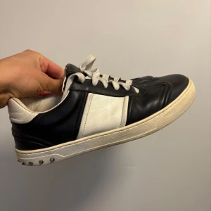 Valentino Garavani svarta och vita sneakers - Säljer ett par Valentino Garavani sneakers i svart och vitt skinn med coola nitar på sulan. Skorna har snörning, rund tå och platt sula. Klassisk design med kontrasterande paneler och vita skosnören. Perfekt för dig som gillar lyxiga detaljer och streetstyle.