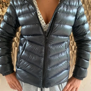 Svart pufferjacka från Colmar - Snygg svart pufferjacka från Colmar med glansig finish och klassisk logga på ärmen. Jackan har horisontella sömmar, dragkedja framtill och huva. Mycket bra skick, dock en liten reva i innerfodralet, se bild. Storlek 152, är som en xs, jag är 15 år, och den har suttit perfekt.