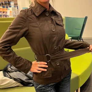 Brun jacka med bälte och fickor - Snygg brun jacka med markerad midja tack vare det breda bältet med metallspänne. Jackan har klassisk krage, stora knappar framtill, bröstficka med lock och två sidfickor. Perfekt för dig som gillar stilren och enkel design.