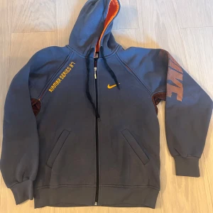 Nike hoodie grå/orange S - Nike hoodie i mörkgrått med orange detaljer och tryck. Hel dragkedja, huva med orange insida och snörning. Snyggt Airmax 90s-tryck på bröstet och stort Nike-tryck på ärmen. Perfekt för dig som gillar sportig streetstyle.