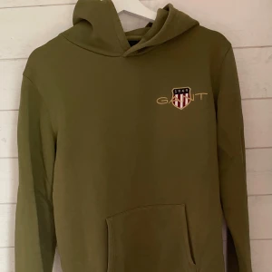 Olivgrön hoodie från Gant - TRYCK INTE PÅ KÖP!                                    Olivgrön hoodie från Gant med broderad logga och sköld på bröstet. Tillverkad i mjuk bomull och har långa ärmar. Är i mycket gott skick och skonsamt använd. Kom gärna med pris förslag. Vid frågor och funderingar fråga:)