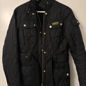 Svart quiltad jacka från Barbour - Svart quiltad jacka från Barbour International med guldiga detaljer, bälte i midjan och flera fickor med tryckknappar. Klassisk design med hög krage och dragkedja framtill. Perfekt för dig som gillar stilren och tidlös look.