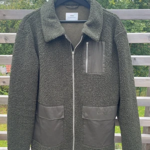 Olivgrön teddyjacka från Ciszere - Säljer en olivgrön teddyjacka från Ciszere i storlek M. Jackan har stora fickor framtill med snygga detaljer i ett annat material och en bröstficka med dragkedja. Perfekt för dig som gillar en chill och trendig look.