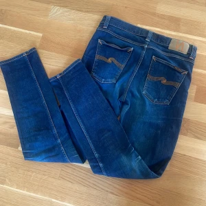 Mörkblå jeans från Nudie Jeans - Säljer ett par mörkblå jeans från Nudie Jeans med klassisk femficksdesign och kontrastsömmar. Jeansen har smal passform och raka ben, samt detaljerade bakfickor med Nudie-loggan. Perfekta för dig som gillar stilrena och tidlösa jeans. pris går att diskutera!