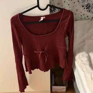 Vinröd långärmad topp från Nelly - Supersöt vinröd topp från Nelly med lång ärm, ribbad struktur och croppad passform. Toppen har en söt knytning framtill och vågig kant nertill för extra detalj. Perfekt för dig som gillar en feminin och trendig look.