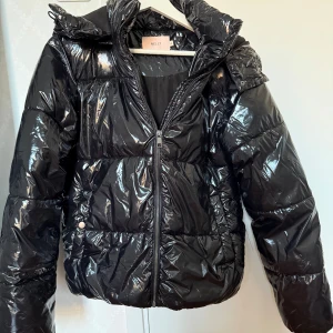 Svart glansig pufferjacka från Nelly - Svart pufferjacka från Nelly i glansigt material med huva och dragkedja framtill. Jackan har quiltad design, långa ärmar och tryckknappar vid fickorna. Perfekt för kalla dagar.