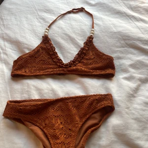 Brun virkad bikini från H&M - Snygg brun bikini från H&M med virkad look och pärldetaljer på axelbanden. Bikinin har ett klassiskt triangelformat överdel och matchande underdel, båda med fina virkade mönster. Perfekt för stranden eller poolen!