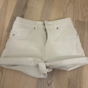 Vita högmidjade jeansshorts - Snygga vita jeansshorts med hög midja och uppvikta ben. Klassisk femficksmodell med bälteshällor och dragkedja framtill. Perfekta för varma dagar och enkel att matcha med allt.
