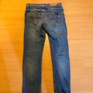 Nudie Jeans blå denim 28/32 - Klassiska blå jeans från Nudie Jeans med orange sömmar och snygga bakfickor med vågig brodyr. Modellen har normal passform och raka ben. Tillverkade i slitstark bomullsdenim, perfekta för en avslappnad stil.