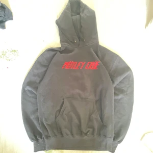 Svart Mötley Crüe hoodie Fruit of the Loom - Svart hoodie från Fruit of the Loom med röd Mötley Crüe-logga på bröstet och stort tryck på ryggen med texten 'Do Fast For Love' och coolt rockmotiv. Klassisk känguruficka och huva. Perfekt för dig som gillar bandmerch och rockstil.