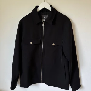 Svart jacka från H&M med dragkedja - Svart overshirt från H&M med klassisk krage, två bröstfickor med knappar och hel dragkedja framtill. Jackan är tillverkad i polyesterblandning och har långa ärmar med knapp vid ärmslut. Enkel och stilren modell som passar till många outfits. 2L1
