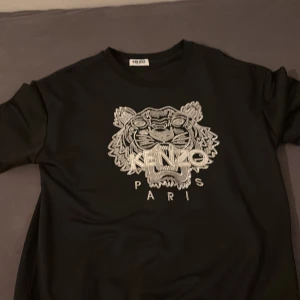 Svart sweatshirt från Kenzo med tiger - Svart sweatshirt från Kenzo med ikoniskt broderat tigerhuvud och Kenzo Paris-text på bröstet. Tröjan har rund halsringning, långa ärmar och är tillverkad i mjuk bomull. Perfekt statement-plagg för dig som gillar streetstyle.