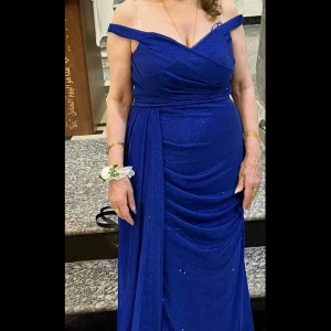 Glittrig lång blå klänning - Säljer en elegant långklänning i djupblå färg med glittriga detaljer. Klänningen har draperad design framtill som ger en snygg siluett och extra volym. Perfekt för dig som vill sticka ut på bal eller fest. Materialet känns lyxigt och skimrar snyggt i ljuset.