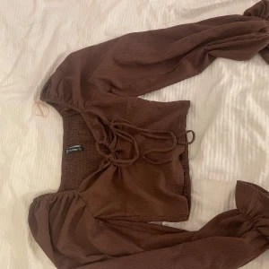 Brun croppad blus med puffärmar - Säljer en brun croppad blus från neyworker med långa puffärmar och snygg snörning framtill. Toppen har en fyrkantig ringning och är gjord i ett lätt, strukturerat tyg som ger en trendig look. Perfekt att styla med höga jeans eller kjol.