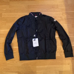 Svart vindjacka från Moncler - Svart vindjacka från Moncler med klassisk ståkrage och dragkedja framtill. Jackan har ribbade muddar, diskret logotyp på ärmen och snygga detaljer i rött och vitt på kragen. Passar perfekt till hösten!