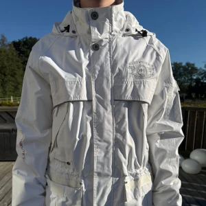 Vit vindjacka från Mountain Horse XS - Snygg vit vindjacka från Mountain Horse med huva, flera fickor och coola logodetaljer på bröstet och ärmen. Jackan har dragsko i midjan och stängs med både dragkedja och knappar. Perfekt för dig som gillar stilren och sportig look. Nypris 800kr