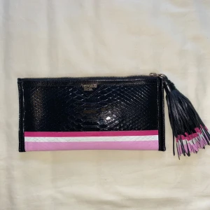 Svart och rosa kuvertväska Victoria's Secret - Snygg kuvertväska från Victoria's Secret i svart och rosa med reptilmönstrad yta. Väskan har en dragkedja upptill och en stor svart tofs med rosa och vita detaljer. Framsidan har en rosa och vit rand längs botten. Perfekt storlek för det viktigaste.