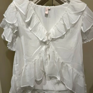 Vit volangblus med knappar - Superfin vit blus med volanger och v-ringning. Blusen har korta ärmar, knappar framtill och är gjord i ett lätt, luftigt material som ger en romantisk vibe. Perfekt för dig som gillar en feminin och trendig look. Aldrig använd 