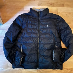 Vändbar pufferjacka från Polo Ralph Lauren - Snygg vändbar pufferjacka från Polo Ralph Lauren med klassisk logga på bröstet. Ena sidan är svart och andra sidan beige. Jackan har hög krage, dragkedja framtill och två fickor på varje sida. Perfekt för dig som vill variera din stil och hålla dig varm.