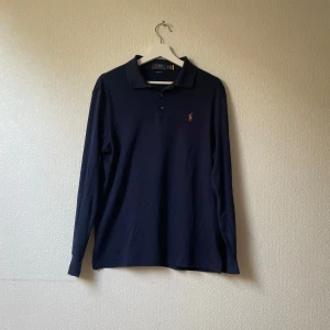 Mörkblå långärmad piké från Ralph Lauren - Klassisk mörkblå långärmad pikétröja från Polo Ralph Lauren med broderad logga på bröstet. Tröjan har krage, knäppning med tre knappar och är i bomull. Perfekt för en clean och tidlös stil.