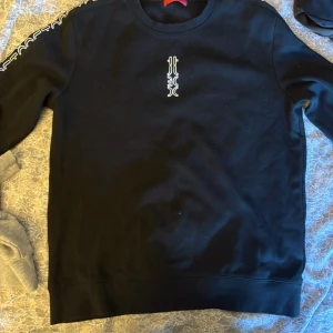 Svart sweatshirt från HUGO - Svart sweatshirt från HUGO med vit logga på bröstet och tryck längs ärmarna. Klassisk rund halsringning och ribbade muddar. Perfekt för en clean och stilren look.