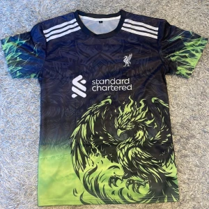 Liverpool fotbollströja grön - Unik Liverpool fotbollströja i polyester med mörkblå och grön färgton, coolt fågelmotiv framtill och vita Adidas-inspirerade ränder på axlarna. Standard Chartered-logga på bröstet och detaljerat mönster i bakgrunden. Perfekt för dig som vill sticka ut på planen.