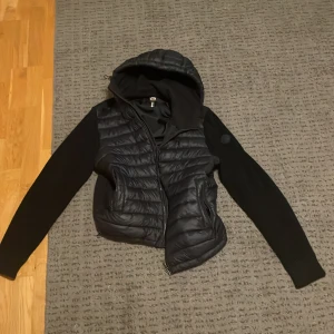 Svart Colmar cardigan med huva - Svart pufferjacka från Colmar med stickade ärmar och vadderad kropp. Jackan har huva, dragkedja framtill och diskret Colmar-logga på ärmen. Perfekt för dig som vill ha en sportig och stilren look under höst och vinter.Det är m men den är ganska stor i storlek jag kan både ha m och l och denna passar mig perfekt men har tröttnat på den,jag är 183 och väger 78kg
