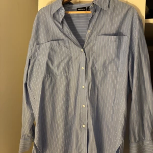 Blå randig skjorta från Pieces - Snygg blå skjorta från Pieces med vita tunna ränder. Klassisk krage, knäppning framtill och två bröstfickor. Långärmad modell i mjuk bomull, perfekt för en clean och stilren look.