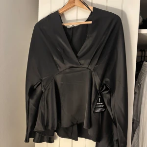 Svart omlottopp i satin - Snygg svart omlottopp i glansigt satinmaterial med v-ringning och långa ärmar. Alla lappar sitter kvar helt oanvänd 