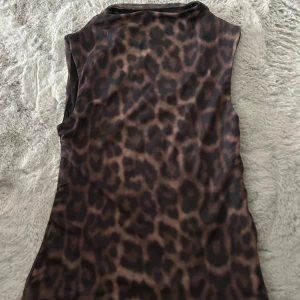 Leopardmönstrad ärmlös topp - Säljer en ärmlös topp med leopardmönster i bruna och svarta toner. Toppen har en mjuk, stretchig känsla och en något högre halsringning. Perfekt för dig som vill sticka ut med ett djurmönster och gillar trendiga detaljer.