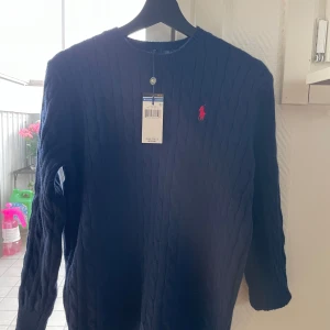 Mörkblå kabelstickad tröja Polo Ralph Lauren - Kabelstickad mörkblå tröja från Polo Ralph Lauren i 100% bomull. Klassisk rund halsringning och långärmad modell med röd broderad logga på bröstet. Perfekt för dig som gillar stilren och tidlös design.