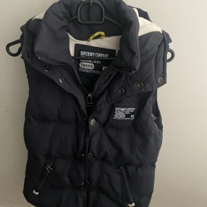 Svart väst från Superdry med luva - Säljer en svart väst från Superdry med luva i sporlek S, modellen är ca 180cm lång. Hör av dig vid funderingar!