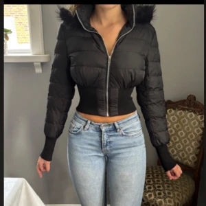 Svart croppad pufferjacka med fuskpäls - Snygg svart croppad pufferjacka med dragkedja och huva med fuskpäls. Jackan har markerad midja, långa ärmar och quiltad design. Perfekt för dig som vill ha en trendig och varm look under kalla dagar.