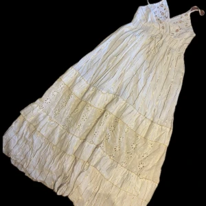 Vit långklänning med broderade detaljer - Romantisk vit långklänning med smala axelband, broderade blommor och volanger. Klänningen har inslag av guldiga ränder, knytning i midjan och vackra spetsdetaljer. Perfekt för sommarens alla tillfällen och ger en bohemisk känsla.