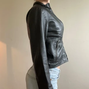 Svart skinnjacka med dragkedja - Snygg svart skinnjacka i kort modell med raka linjer och tight passform. Jackan har långa ärmar, dragkedja framtill och vid fickorna. Perfekt för dig som vill ha en klassisk och stilren look. Liten i storleken (164) men passar en xxs eller 32 tänker jag.