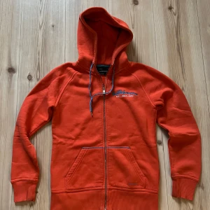Röd/orange hoodie från Peak Performance S - Snygg röd hoodie från Peak Performance med blå snören i huvan och dragkedja framtill. Broderad logga på bröstet och praktiska fickor. Perfekt för chill dagar eller när du vill vara extra bekväm.