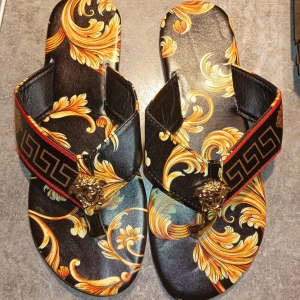 Svarta sandaler med guldmönster - Unika sandaler med svart bas och detaljerat guld- och orange barockmönster. Bred rem med grekiskt mönster i svart och guld samt röd kant. Snygg guldfärgad Medusa-detalj framtill. Perfekta för dig som vill sticka ut med en lyxig vibe.