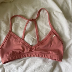 Rosa sportig linnetopp från Gymshark - Säljer en snygg rosa linnetopp från Gymshark med smala korsade axelband i ryggen och diskret logga framtill. Toppen är i mjukt och stretchigt material, perfekt för träning eller chill. Enkel och stilren design med V-ringning.