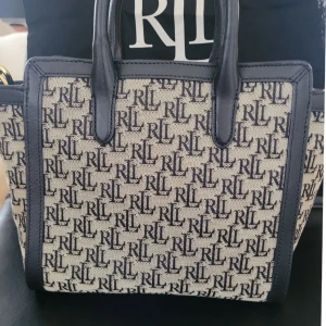 Ralph Lauren RL monogram handväska - Snygg handväska från Lauren Ralph Lauren med RL-monogram över hela väskan. Väskan är beige med svarta detaljer och svarta handtag i skinn. Guldiga metalldetaljer och dragkedja. Perfekt storlek för vardagens essentials och har både axelrem och handtag.