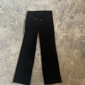 Svarta velourbyxor Juicy Couture S - Svarta mjukisbyxor från Juicy Couture i mjuk velour med bred resårmidja och snörning framtill. Byxorna har raka ben, fickor fram och bak med snygga silverknappar. Dom är i bra skick bara lite noppriga innanför vid midjan men inget man tänker på! Annars är dom helt felfria🩵(Nypris 1200 kr)