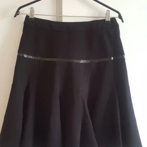 Svart kortkjol med glansig detalj - Svart kortkjol med utsvängd form och en glansig svart banddetalj runt om. Kjolen har en synlig dragkedja bak och är tillverkad i ett tjockare, mjukt material som ger en snygg siluett. Perfekt för dig som gillar stilrena plagg med en twist.