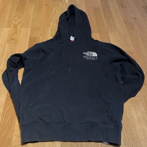 Svart hoodie The North Face Denali S - Svart hoodie från The North Face med Denali-tryck på bröstet och stor grafisk print på ryggen. Klassisk känguruficka, snörning i huvan och ribbade muddar. Perfekt för dig som gillar streetwear och outdoor-vibe. lite sliten där av priset.