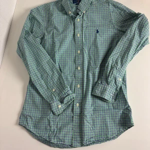 Rutig skjorta från Ralph Lauren M - Snygg rutig skjorta från Ralph Lauren i grönt, blått och vitt. Slim fit-modell med klassisk krage, knappar framtill och broderad logga på bröstet. Perfekt för dig som gillar preppy stil och vill sticka ut med färg. Tillverkad i mjuk bomull.