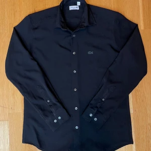Svart skjorta från Lacoste  - Stilren svart skjorta från Lacoste i slim fit-modell. Skjortan har klassisk krage, knäppning framtill och diskret Lacoste-logga broderad på bröstet. Långärmad och tillverkad i mjukt bomullstyg som känns skönt mot huden.