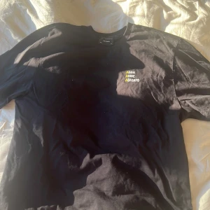 Svart t-shirt Axel Arigato  - Svart t-shirt från Axel Arigato i 100% bomull. Klassisk passform med korta ärmar och rund hals. På bröstet finns broderad text i vitt och gult: 'ADEN ASME ARIGATO' med tre gula A. Perfekt för dig som gillar stilrena streetwear-plagg.