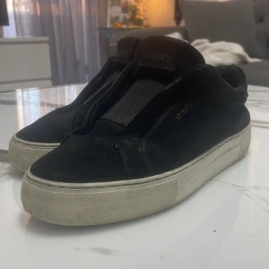 Svarta sneakers från Axel Arigato - Svarta sneakers från Axel Arigato med minimalistisk design och vit platt sula. Skorna är i skinn och har en stilren look utan synlig snörning, perfekt för dig som gillar cleana vibes. Märkeslogga syns på sidan och sulan.