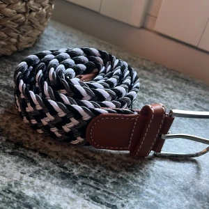 Flätat svartvitt bälte med läderdetalj - Snyggt flätat bälte i svart, vitt och grått med bruna läderdetaljer vid spännet. Bältet har ett coolt mönster och är gjort av syntetmaterial med en klassisk metallspänne. Perfekt för att ge outfiten lite extra edge.