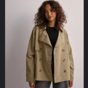 Beige kort trenchcoat med knappar - Stilren beige trenchcoat med dubbelknäppning och bred krage. Jackan har en loose passform och är tillverkad i ett slätt material. Perfekt för dig som gillar klassiska och tidlösa plagg med modern twist.
