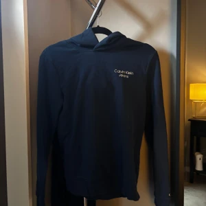 Mörkblå hoodie från Calvin Klein Jeans - Snygg mörkblå hoodie från Calvin Klein Jeans med diskret logga på bröstet. Klassisk modell med huva och långa ärmar, perfekt för en chill och stilren look. Tillverkad i mjuk bomull för extra komfort.