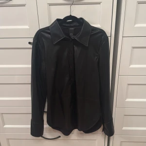 Svart skinnskjorta från Lindex XS - Snygg svart skjorta i skinnimitation från Lindex. Klassisk krage, långa ärmar och knäppning framtill. Perfekt för dig som vill ha en edgy look med stilren känsla. Skjortan har en rak passform och är tillverkad i mjukt syntetmaterial.